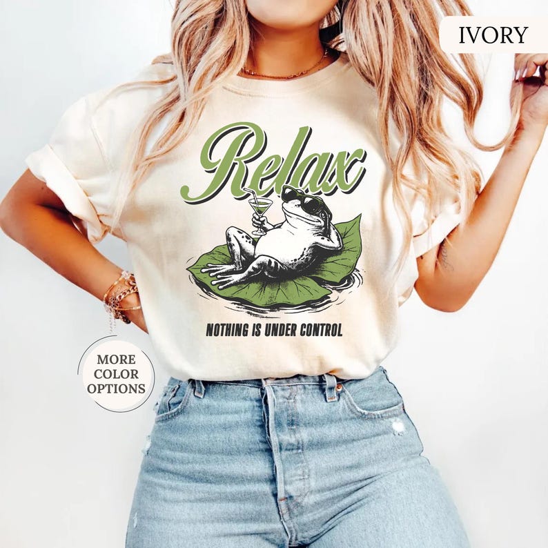 May include: Ivory-colored t-shirt featuring a relaxed frog graphic. The frog, wearing sunglasses and holding a cocktail, is lounging on a lily pad. The shirt has the word "Relax" above the frog and "Nothing is Under Control" below.