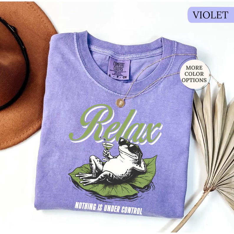 May include: A violet Comfort Colors t-shirt with a graphic of a frog wearing sunglasses and holding a cocktail, lounging on a lily pad. The word "Relax" is above the frog, and "Nothing is Under Control" is below. A gold necklace is draped on the shirt.