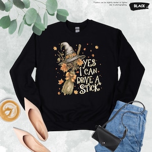 Op de afbeelding: Zwarte sweatshirt met een heksenhoed, bezem en herfstbladeren grafisch ontwerp. De tekst "YES I CAN DRIVE A STICK" is in het wit gedrukt. De sweatshirt is een crew neck stijl.