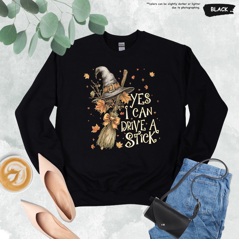 Op de afbeelding: Zwarte sweatshirt met een heksenhoed, bezem en herfstbladeren grafisch ontwerp. De tekst "YES I CAN DRIVE A STICK" is in het wit gedrukt. De sweatshirt is een crew neck stijl.