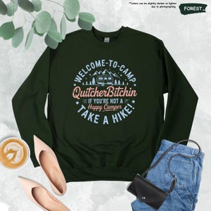 May include: A dark green sweatshirt with the text "WELCOME-TO-CAMP QuitcherBitchin IF YOU'RE NOT A Happy Camper TAKE A HIKE!" in white, pink, and blue. The design includes a camper, mountains, and trees.