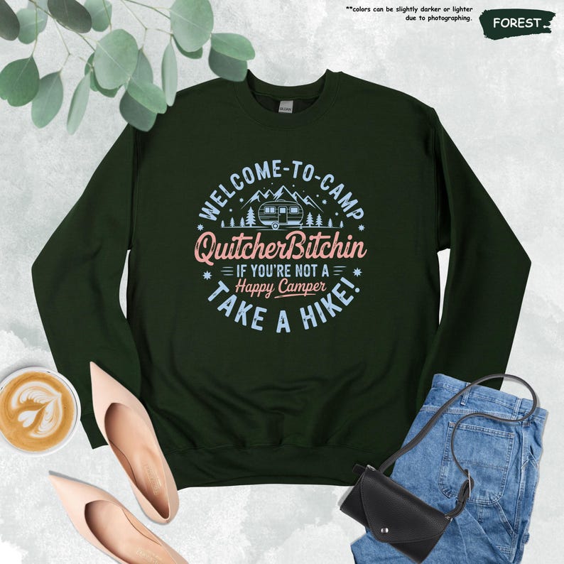 May include: A dark green sweatshirt with the text "WELCOME-TO-CAMP QuitcherBitchin IF YOU'RE NOT A Happy Camper TAKE A HIKE!" in white, pink, and blue. The design includes a camper, mountains, and trees.