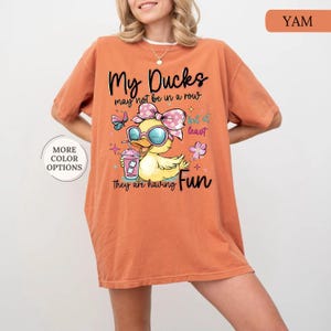 May include: Orange t-shirt with a graphic of a yellow duck wearing sunglasses and a bow, holding a drink. Text on the shirt reads "My Ducks may not be in a row but at least they are having Fun." The shirt also has a "More Color Options" button.