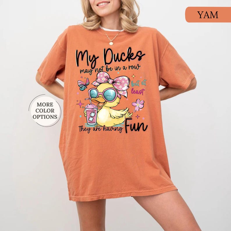 May include: Orange t-shirt with a graphic of a yellow duck wearing sunglasses and a bow, holding a drink. Text on the shirt reads "My Ducks may not be in a row but at least they are having Fun." The shirt also has a "More Color Options" button.