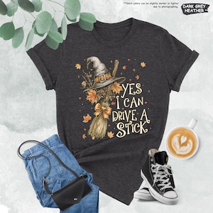 Pode incluir: T-shirt cinza escuro mesclado com um chap&eacute;u de bruxa, vassoura e folhas de outono. O texto "YES I CAN DRIVE A STICK" est&aacute; impresso na camiseta. A imagem inclui jeans azuis, uma bolsa preta e t&ecirc;nis pretos de cano alto.