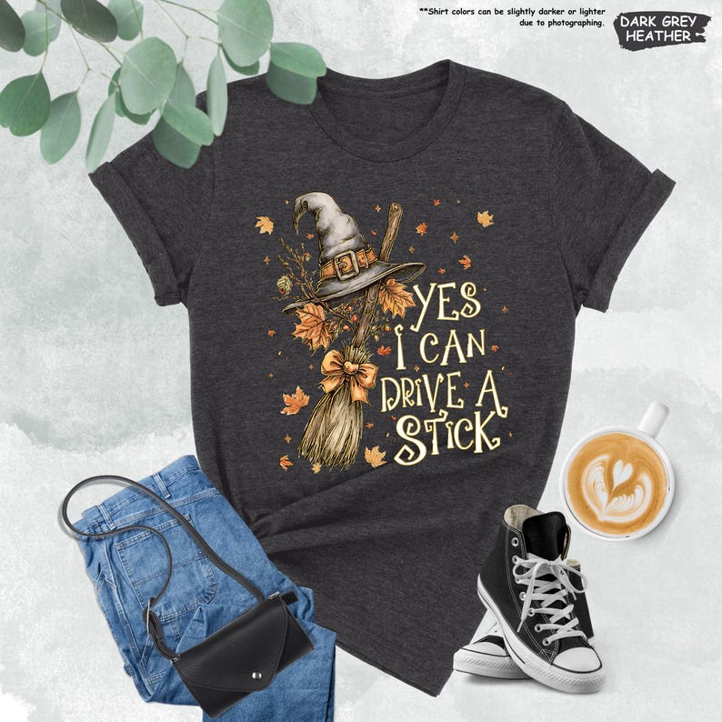 Pode incluir: T-shirt cinza escuro mesclado com um chap&eacute;u de bruxa, vassoura e folhas de outono. O texto "YES I CAN DRIVE A STICK" est&aacute; impresso na camiseta. A imagem inclui jeans azuis, uma bolsa preta e t&ecirc;nis pretos de cano alto.