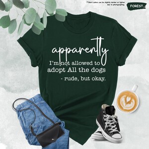 May include: Forest green t-shirt with the text "apparently I'm not allowed to adopt All the dogs - rude, but okay." The shirt is paired with blue jeans, a black crossbody bag, and black high-top sneakers. A cup of coffee with latte art is also visible.