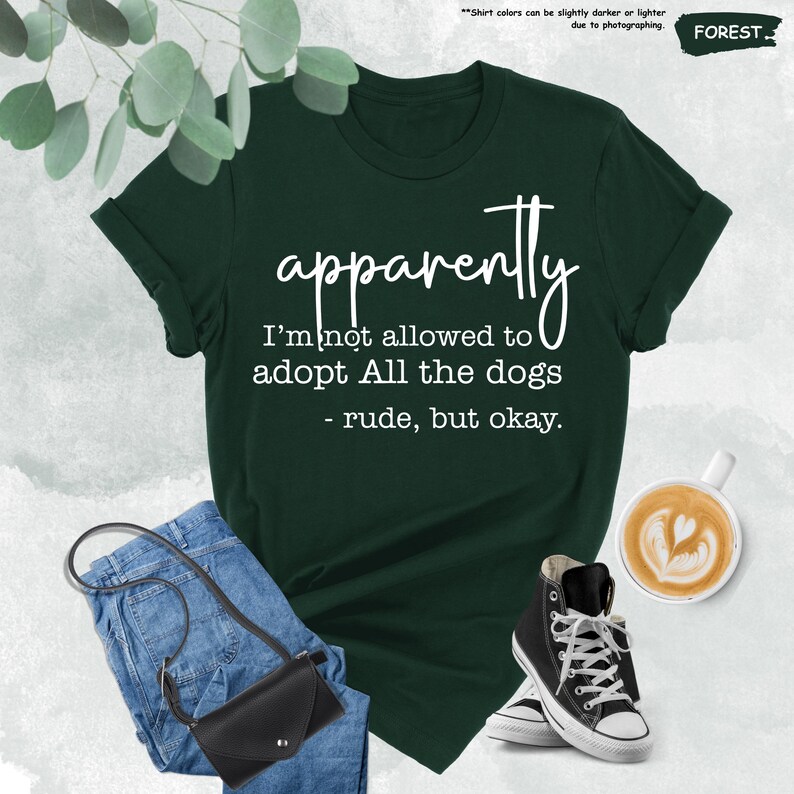 May include: Forest green t-shirt with the text "apparently I'm not allowed to adopt All the dogs - rude, but okay." The shirt is paired with blue jeans, a black crossbody bag, and black high-top sneakers. A cup of coffee with latte art is also visible.
