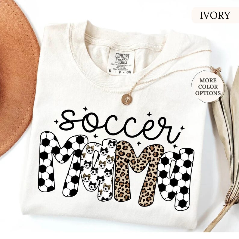 Pu&ograve; includere: T-shirt color avorio con le parole "soccer mama" in corsivo nero e lettere maiuscole. La parola "mama" presenta motivi a pallone da calcio, stampa leopardata e motivi a pallone da calcio con fiocchi. Una collana d'oro &egrave; drappeggiata sulla maglietta.