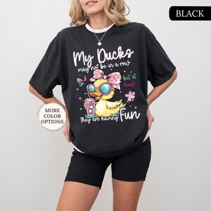 May include: Black t-shirt with the text "My Ducks may not be in a row but at least they are having Fun." The graphic features a cartoon duck wearing sunglasses and a bow, holding a drink. The shirt also has the words "More Color Options" on it.
