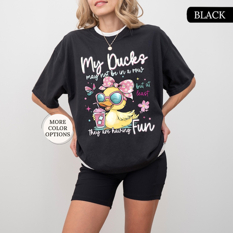 May include: Black t-shirt with the text "My Ducks may not be in a row but at least they are having Fun." The graphic features a cartoon duck wearing sunglasses and a bow, holding a drink. The shirt also has the words "More Color Options" on it.