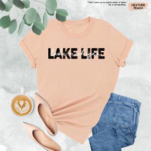 May include: A heather peach-colored t-shirt with the words "LAKE LIFE" printed in black and white. The shirt is paired with light blue jeans and beige flats. A cup of coffee with latte art is also visible.