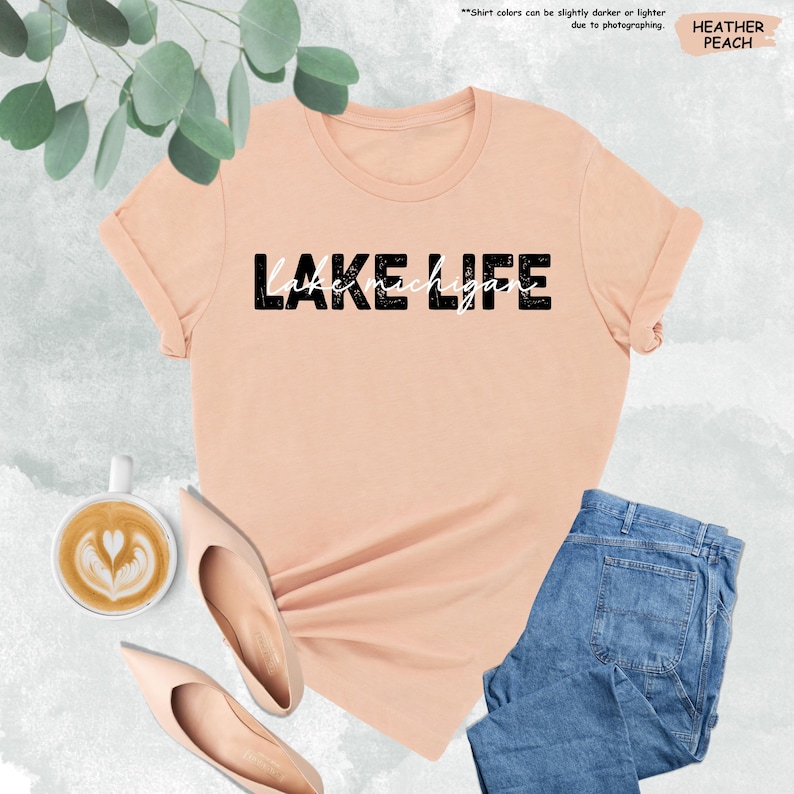 May include: A heather peach-colored t-shirt with the words "LAKE LIFE" printed in black and white. The shirt is paired with light blue jeans and beige flats. A cup of coffee with latte art is also visible.