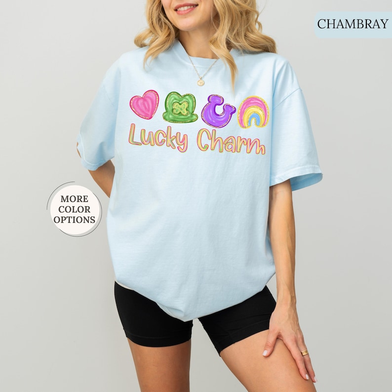 May include: Light blue t-shirt with the text "Lucky Charm" and colorful graphics of a heart, shamrock, horseshoe, and rainbow. The shirt has short sleeves and a crew neck.