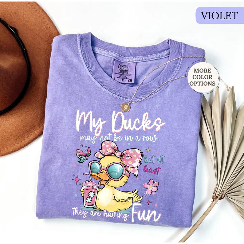 May include: Violet Comfort Colors t-shirt with the text "My Ducks may not be in a row but at least they are having Fun." The shirt features a cartoon duck wearing sunglasses and a bow, holding a drink.