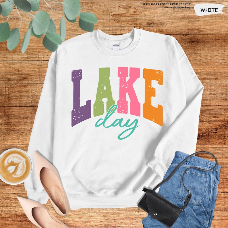 Op de afbeelding: Witte sweatshirt met de tekst "LAKE day" in paars, groen, roze, oranje en turquoise. De sweatshirt wordt gecombineerd met een blauwe jeans, een zwarte tas en nude schoenen. Een kopje koffie is ook zichtbaar.