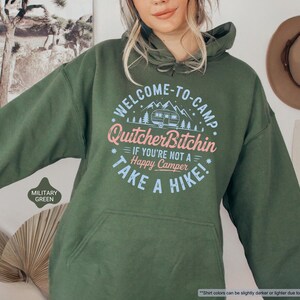 May include: Military green hoodie with a front pocket and drawstring hood. The front features a circular graphic with the text "WELCOME-TO-CAMP Quitcher Bitchin IF YOU'RE NOT A Happy Camper TAKE A HIKE!" in pink and blue.
