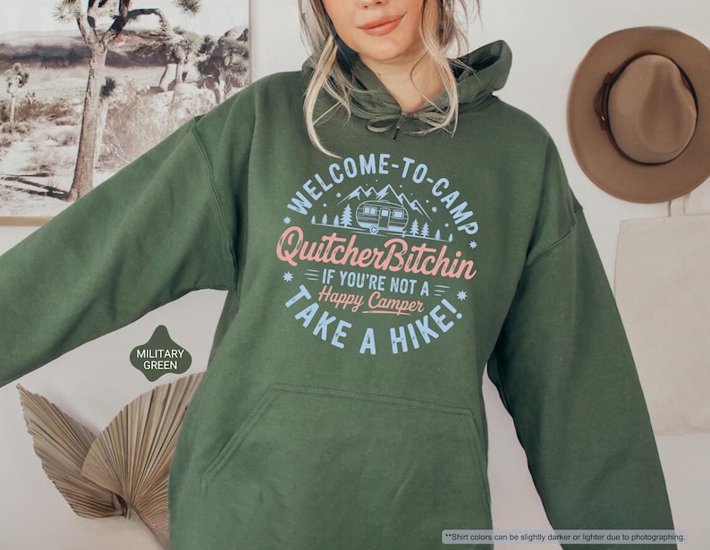 May include: Military green hoodie with a front pocket and drawstring hood. The front features a circular graphic with the text "WELCOME-TO-CAMP Quitcher Bitchin IF YOU'RE NOT A Happy Camper TAKE A HIKE!" in pink and blue.