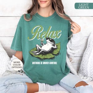 May include: A light green t-shirt with the word "Relax" in a script font above a cartoon frog wearing sunglasses and holding a cocktail. The text "NOTHING IS UNDER CONTROL" is below the frog.