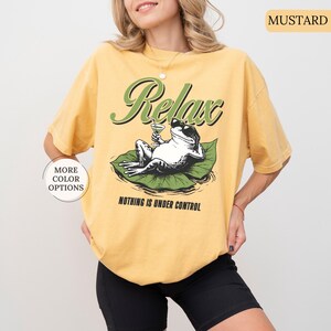 May include: Mustard-colored t-shirt with a graphic of a frog wearing sunglasses, holding a cocktail, and lounging on a lily pad. The shirt has the words "Relax" and "Nothing is Under Control" printed on it.