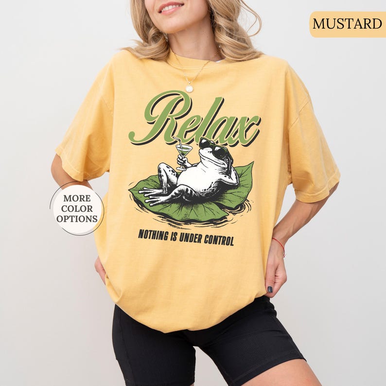 May include: Mustard-colored t-shirt with a graphic of a frog wearing sunglasses, holding a cocktail, and lounging on a lily pad. The shirt has the words "Relax" and "Nothing is Under Control" printed on it.