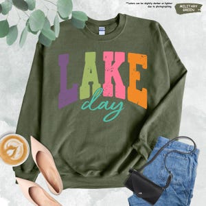 Op de afbeelding: Militairgroen sweatshirt met de tekst "LAKE day" in paarse, groene, roze en oranje blokletters, met een turquoise "day"-script. Het sweatshirt is gestyled met een jeans, een zwarte handtas en nude hakken.