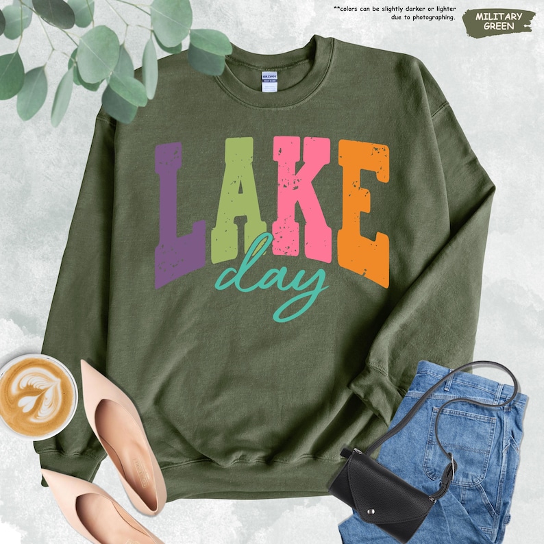 Op de afbeelding: Militairgroen sweatshirt met de tekst "LAKE day" in paarse, groene, roze en oranje blokletters, met een turquoise "day"-script. Het sweatshirt is gestyled met een jeans, een zwarte handtas en nude hakken.