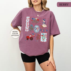 May include: Berry-colored t-shirt with a collage of patriotic-themed illustrations. The design includes a red tractor with an American flag, a goose wearing a hat, floral patterns, and the text "let freedom ring."