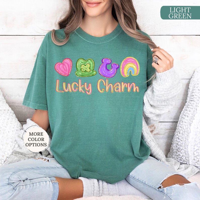May include: A light green t-shirt with the words "Lucky Charm" in a colorful font. Above the text are four colorful graphics: a pink heart, a green shamrock, a purple horseshoe, and a rainbow.