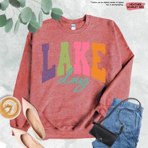 Op de afbeelding: Heather scharlakenrood sweatshirt met de tekst "LAKE day" in paars, groen, roze, oranje en turquoise. De afbeelding bevat een paar beige hakken, een kopje koffie, een zwarte handtas en een spijkerbroek.