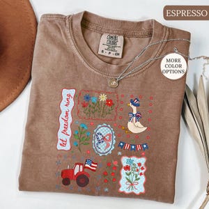 May include: Espresso-colored Comfort Colors t-shirt with patriotic-themed embroidered patches. Patches include a goose in a hat, flowers, a tractor, and the text "let freedom ring". A silver necklace is draped across the shirt.