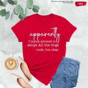 May include: A red t-shirt with the text "apparently I'm not allowed to adopt All the dogs - rude, but okay." The shirt is paired with blue jeans, beige heels, and a cup of coffee with latte art.