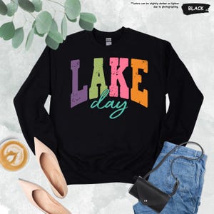 Op de afbeelding: Zwart sweatshirt met de tekst "LAKE day" in paarse, groene, roze en oranje blokletters. Het woord "day" is in een turquoise scriptlettertype. Het sweatshirt wordt gecombineerd met een jeans, schoenen en een zwarte tas.