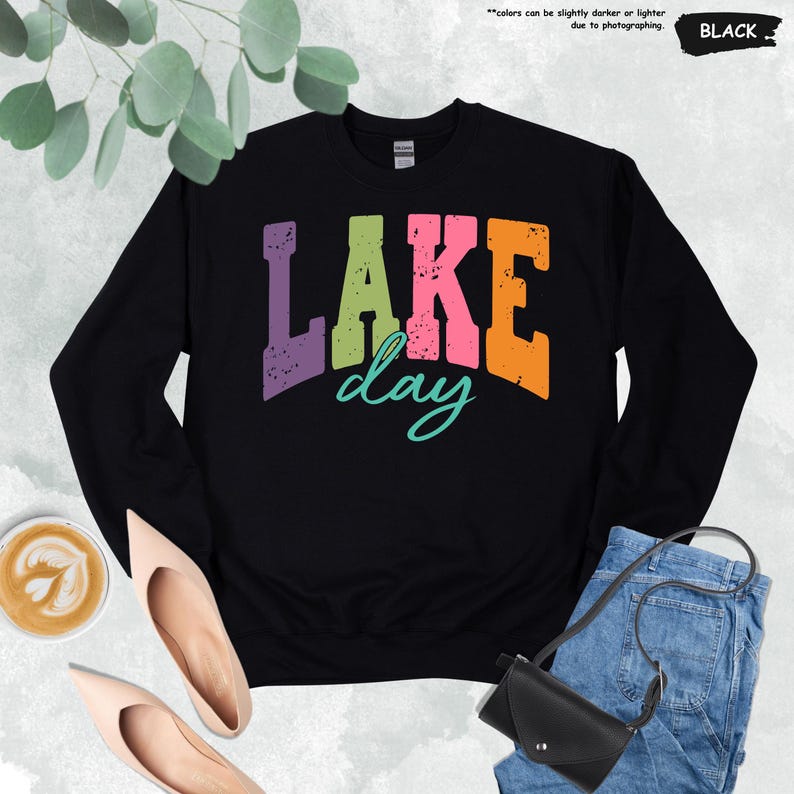 Op de afbeelding: Zwart sweatshirt met de tekst "LAKE day" in paarse, groene, roze en oranje blokletters. Het woord "day" is in een turquoise scriptlettertype. Het sweatshirt wordt gecombineerd met een jeans, schoenen en een zwarte tas.