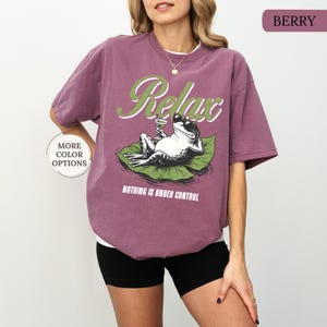 May include: Berry-colored t-shirt with a graphic of a frog relaxing on a lily pad, holding a cocktail. The word "Relax" is arched above the frog, with the phrase "Nothing is Under Control" below.