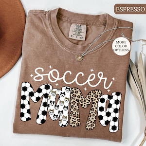 Pu&ograve; includere: T-shirt color espresso con le parole "soccer mama" in scrittura bianca. La parola "mama" &egrave; disegnata con palloni da calcio, stampa leopardata e motivi a pallone da calcio. La maglietta ha un girocollo e maniche corte. Una collana d'argento &egrave; drappeggiata sulla maglietta.