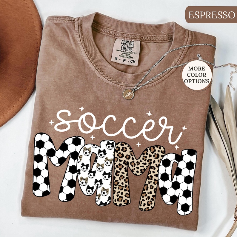 Pu&ograve; includere: T-shirt color espresso con le parole "soccer mama" in scrittura bianca. La parola "mama" &egrave; disegnata con palloni da calcio, stampa leopardata e motivi a pallone da calcio. La maglietta ha un girocollo e maniche corte. Una collana d'argento &egrave; drappeggiata sulla maglietta.
