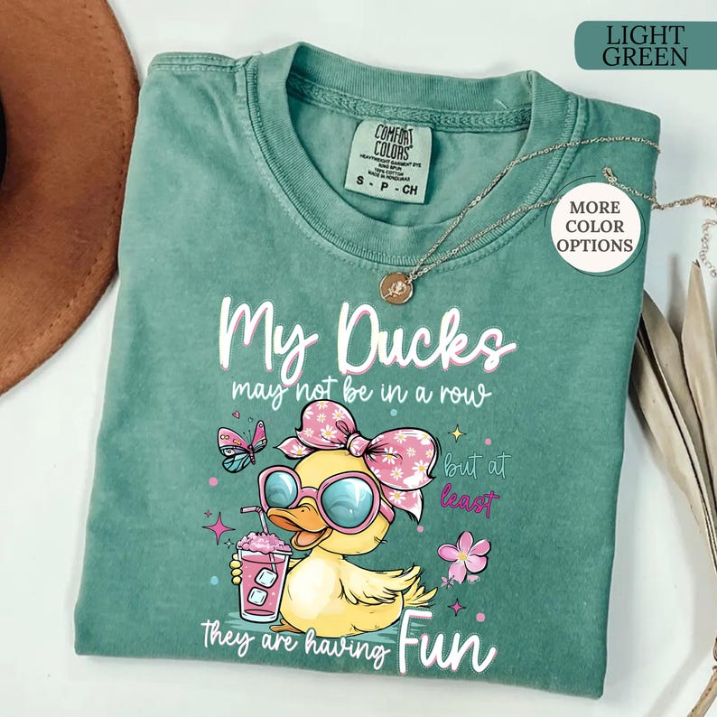 May include: Light green Comfort Colors t-shirt with the text "My Ducks may not be in a row but at least they are having Fun." Features a cartoon duck wearing sunglasses and a pink bow, holding a drink.