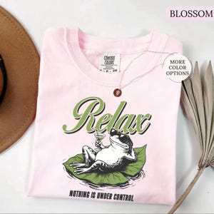 May include: A light pink t-shirt with the word "Relax" in green script and a graphic of a frog wearing sunglasses and holding a cocktail. The phrase "Nothing is Under Control" is printed below the frog. The shirt has a crew neck and short sleeves.