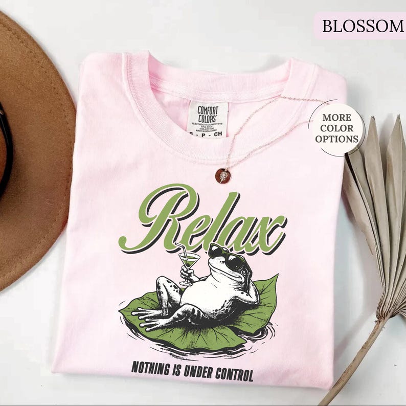 May include: A light pink t-shirt with the word "Relax" in green script and a graphic of a frog wearing sunglasses and holding a cocktail. The phrase "Nothing is Under Control" is printed below the frog. The shirt has a crew neck and short sleeves.
