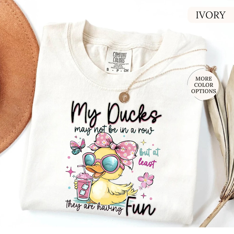 May include: Ivory-colored t-shirt with the text "My Ducks may not be in a row but at least they are having Fun." The graphic features a cartoon duck wearing sunglasses and a bow, holding a drink. A butterfly and flowers are also included.
