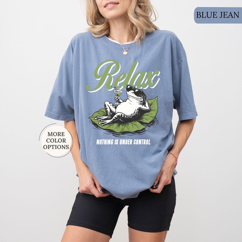 May include: Blue jean colored t-shirt with a graphic of a frog wearing sunglasses, relaxing on a lily pad and holding a cocktail. The word "Relax" is above the frog in green script, and the phrase "Nothing is Under Control" is below.
