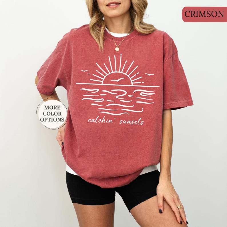 May include: Crimson-colored t-shirt with a white graphic of a sunset over the ocean, featuring the text "catchin' sunsets." The shirt has short sleeves and a relaxed fit. The words "More Color Options" are also visible.