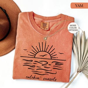 May include: A yam-colored Comfort Colors t-shirt with a black graphic of a sunset over the ocean, with the words "catchin' sunsets" below. A gold necklace with a pendant is on the shirt. The shirt is next to a brown hat and a dried plant.