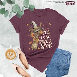 Pode incluir: Camiseta bord&ocirc; com chap&eacute;u de bruxa, vassoura e o texto "YES I CAN DRIVE A STICK". O design inclui folhas de outono e um la&ccedil;o. A camiseta &eacute; combinada com jeans azuis e sapatos nude.