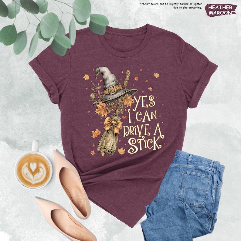 Pode incluir: Camiseta bord&ocirc; com chap&eacute;u de bruxa, vassoura e o texto "YES I CAN DRIVE A STICK". O design inclui folhas de outono e um la&ccedil;o. A camiseta &eacute; combinada com jeans azuis e sapatos nude.