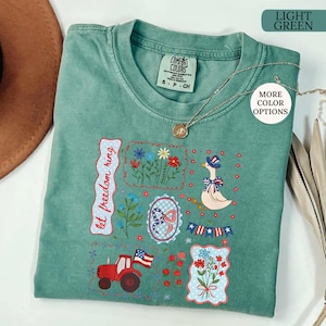 May include: Light green Comfort Colors t-shirt with patriotic embroidery. Features a red tractor, a goose in a patriotic hat, and floral designs. The text "let freedom ring" is also embroidered on the shirt. A silver necklace is draped across the shirt.