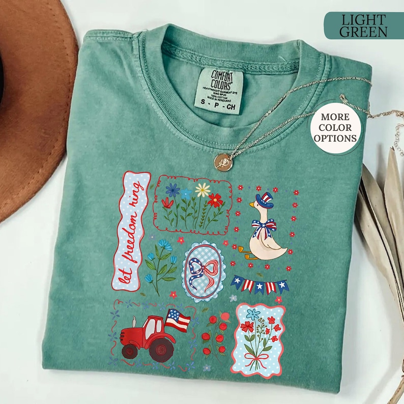 May include: Light green Comfort Colors t-shirt with patriotic embroidery. Features a red tractor, a goose in a patriotic hat, and floral designs. The text "let freedom ring" is also embroidered on the shirt. A silver necklace is draped across the shirt.