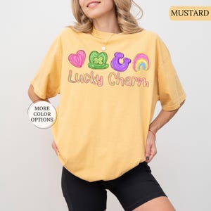 May include: Mustard-colored t-shirt with the text "Lucky Charm" and colorful graphic icons. The icons include a pink heart, green shamrock, purple horseshoe, and a rainbow. The shirt has a relaxed fit and is worn with black shorts.