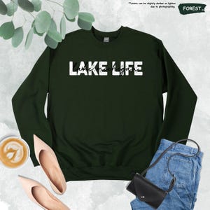 Peut inclure: Un sweat-shirt &agrave; col rond vert for&ecirc;t arborant le texte blanc vieilli LAKE LIFE avec le texte cursif Lake Michigan superpos&eacute;. Le v&ecirc;tement pr&eacute;sente des manches longues et un col c&ocirc;tel&eacute;, pr&eacute;sent&eacute; comme un article vestimentaire &agrave; plat.
